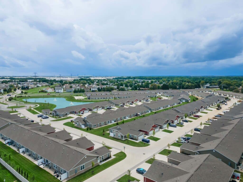 Aerial view of a suburban neighborhood with rows of single-story houses, well-maintained lawns, and a small pond in the background under a cloudy sky.