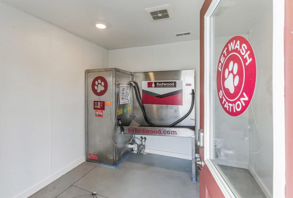 A self-service pet wash station with a metal wash tub, hoses, and labeled instructions, viewed through a door with "Pet Wash Station" signage.