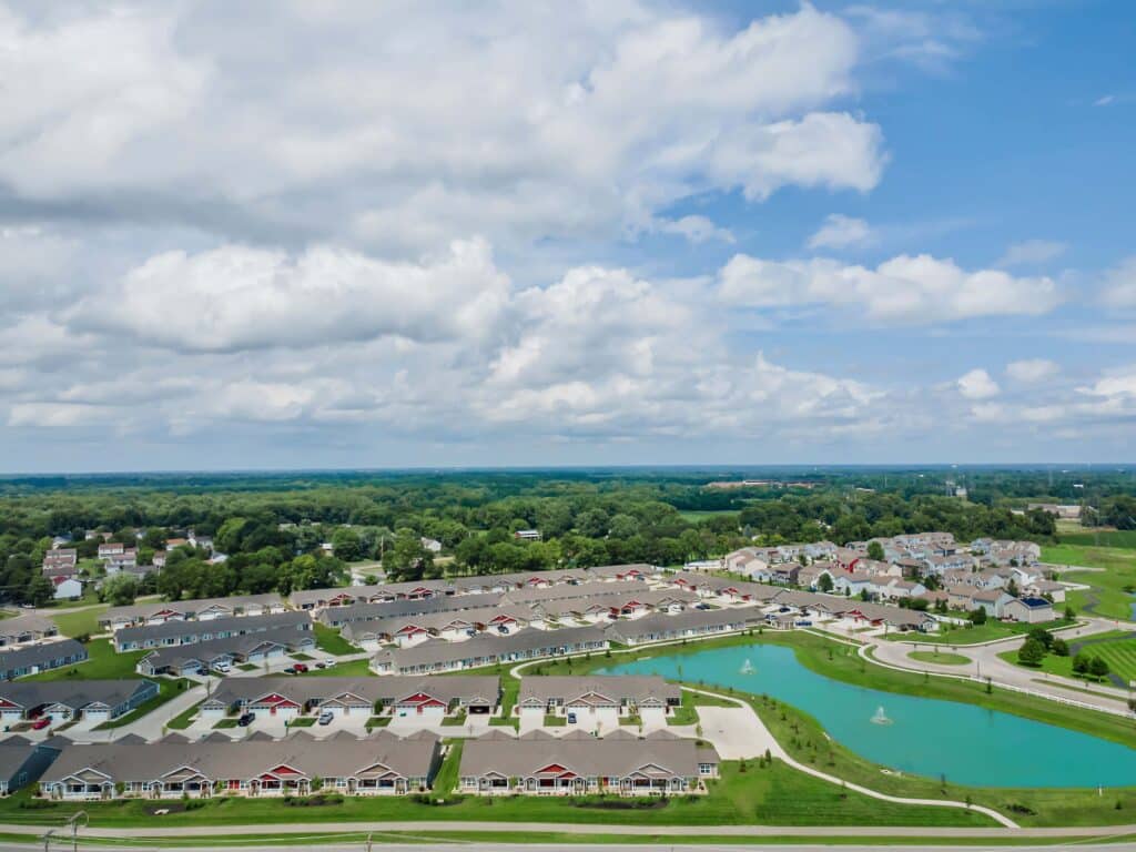 Aerial view of a suburban neighborhood with rows of houses, green lawns, small lakes, and fountains under a partly cloudy sky.