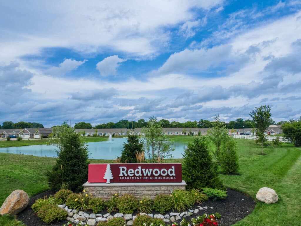 A landscaped area with a “Redwood Apartment Neighborhoods” sign in front of a pond, townhouses, and a partly cloudy sky.