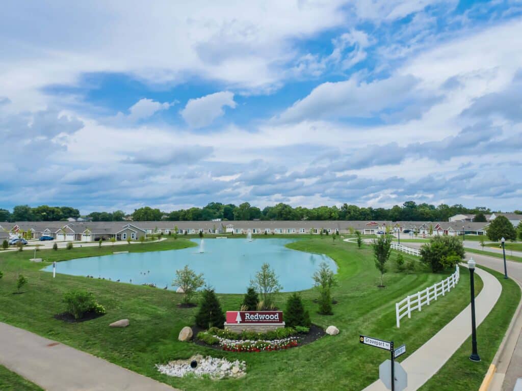 A landscaped neighborhood pond with a fountain, surrounded by grass, houses, trees, and a white fence, under a partly cloudy sky. A sign reads "Redwood.