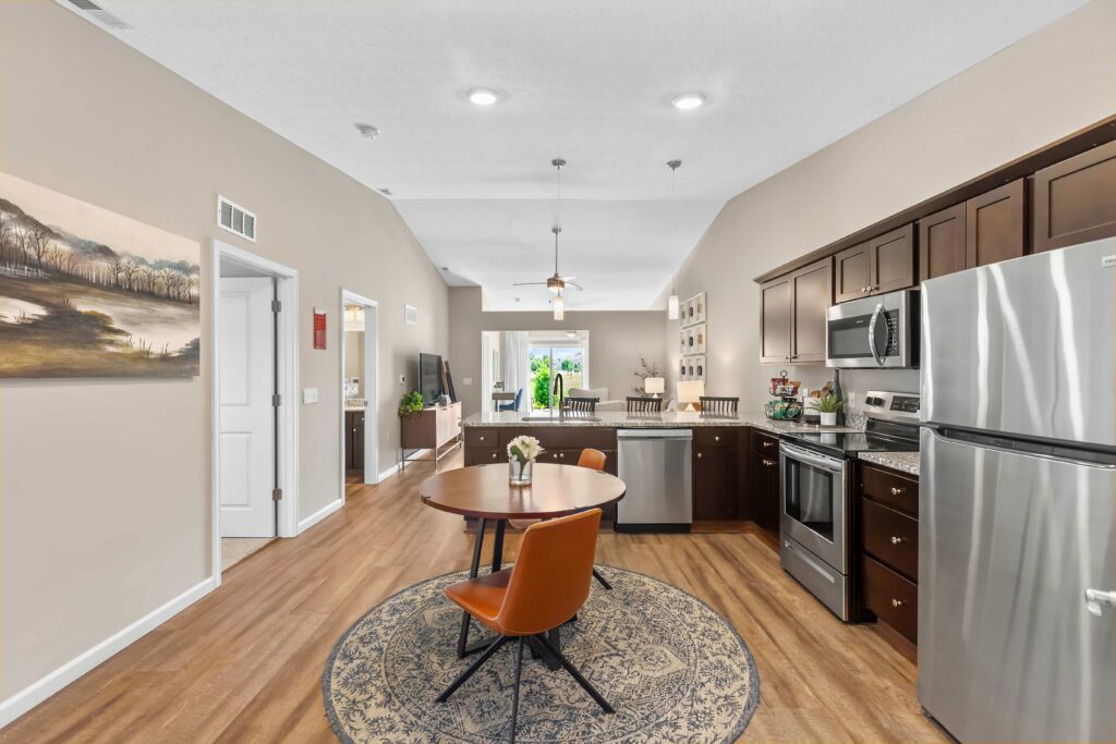 Modern open-concept kitchen and dining area with stainless steel appliances, dark wood cabinets, round table, and two chairs on a rug, leading to a bright living space.