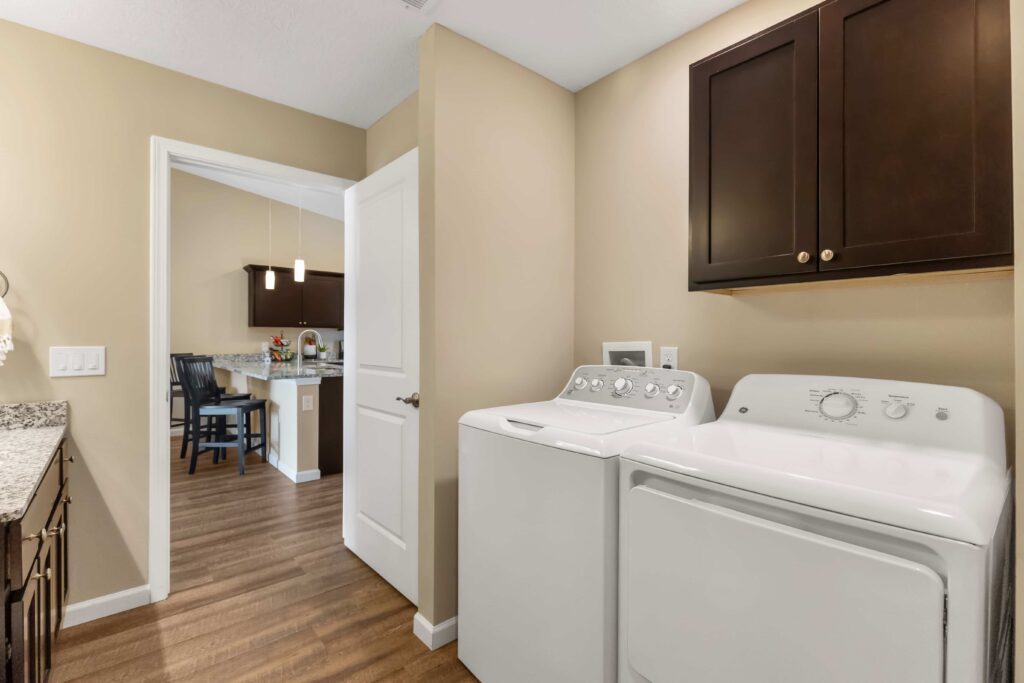 Laundry room with a washing machine and dryer next to a cabinet, viewed from the open doorway leading to a kitchen and dining area.