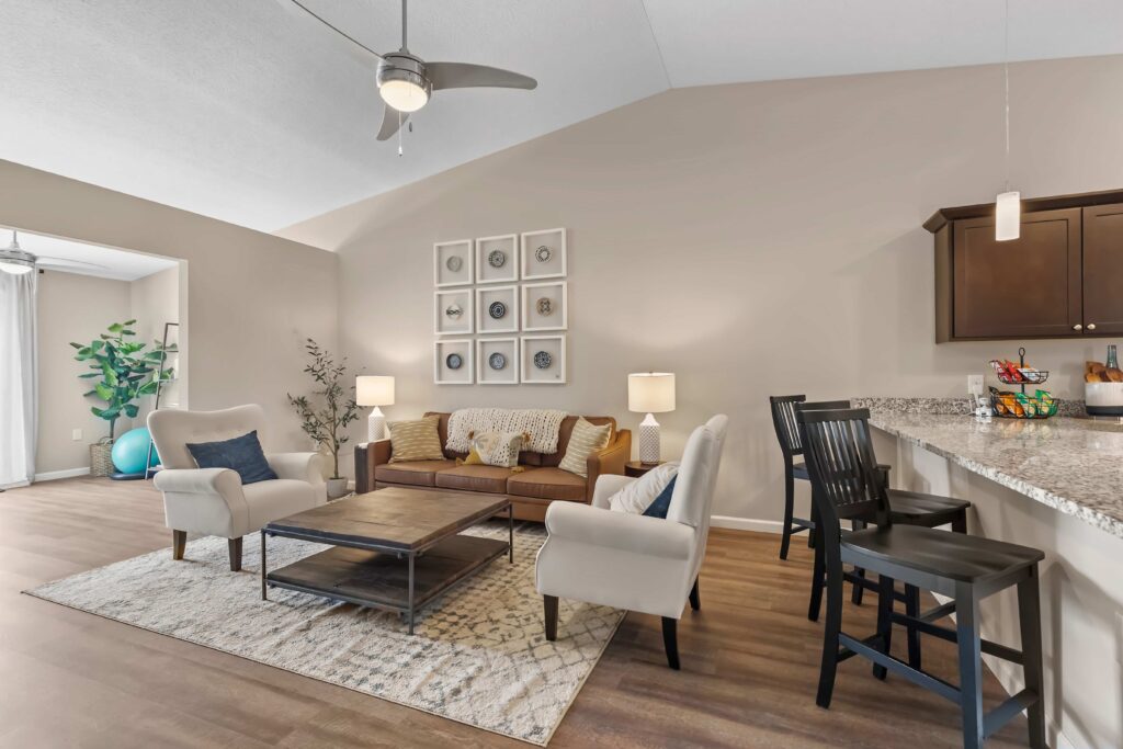 A modern living room with a brown leather sofa, two armchairs, a wooden coffee table, a kitchen bar with stools, and neutral decor under high ceilings.