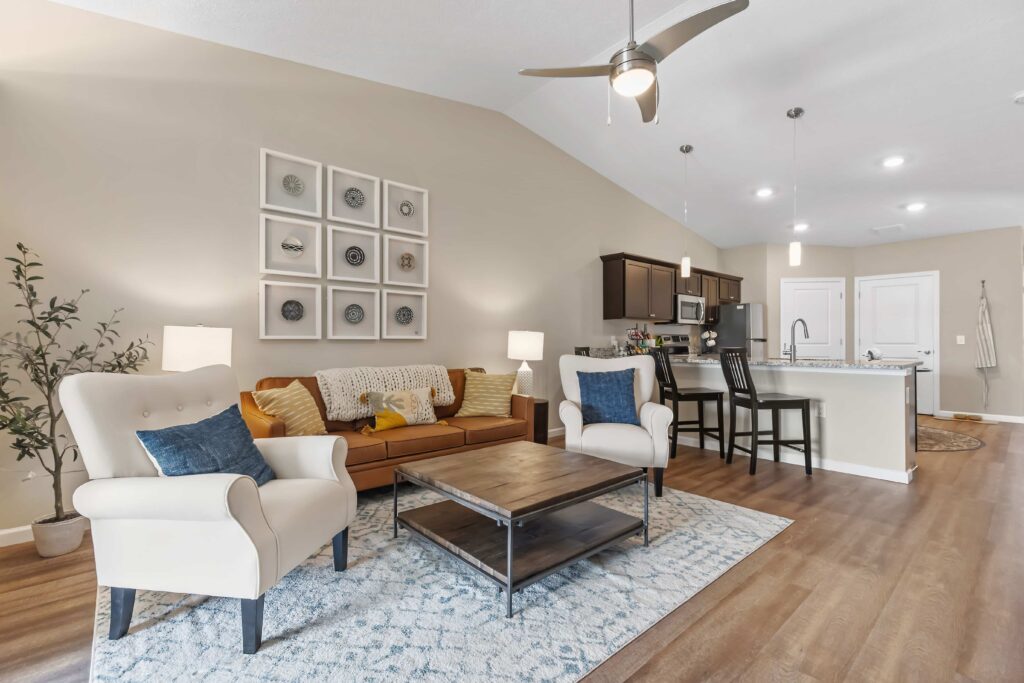 Open-concept living room and kitchen with neutral tones, modern furnishings, a brown sofa, two beige armchairs, a rug, and wall art above the couch.