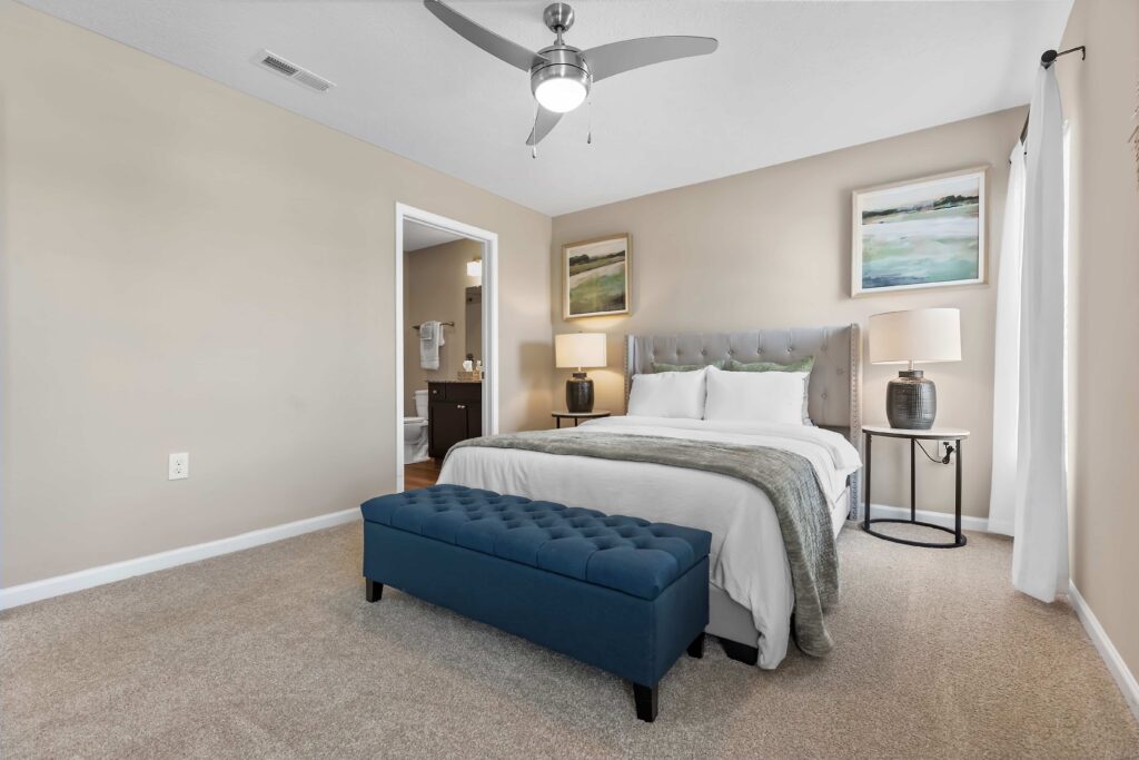A bedroom with a neatly made bed, two bedside tables with lamps, a blue upholstered bench at the foot of the bed, and an open door to a bathroom.