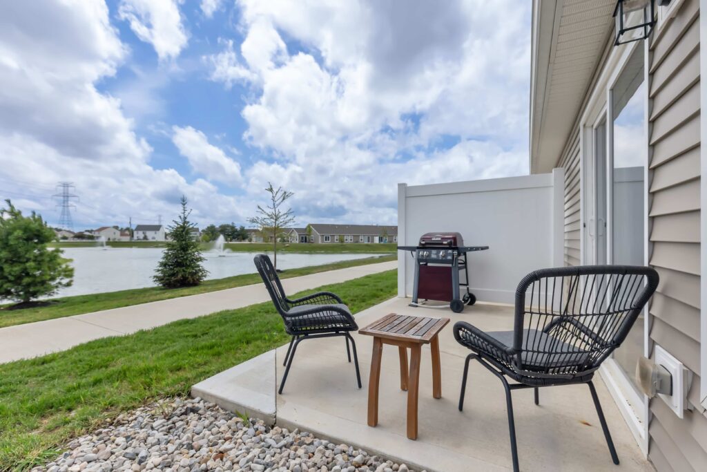 Small patio with two black chairs, a wooden table, and a grill overlooks a pond and neighborhood under a partly cloudy sky.