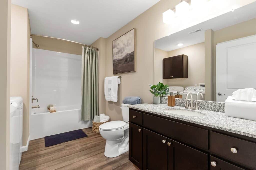 Modern bathroom with a granite countertop, dark wood cabinets, toilet, bathtub with shower curtain, and neutral wall decor.