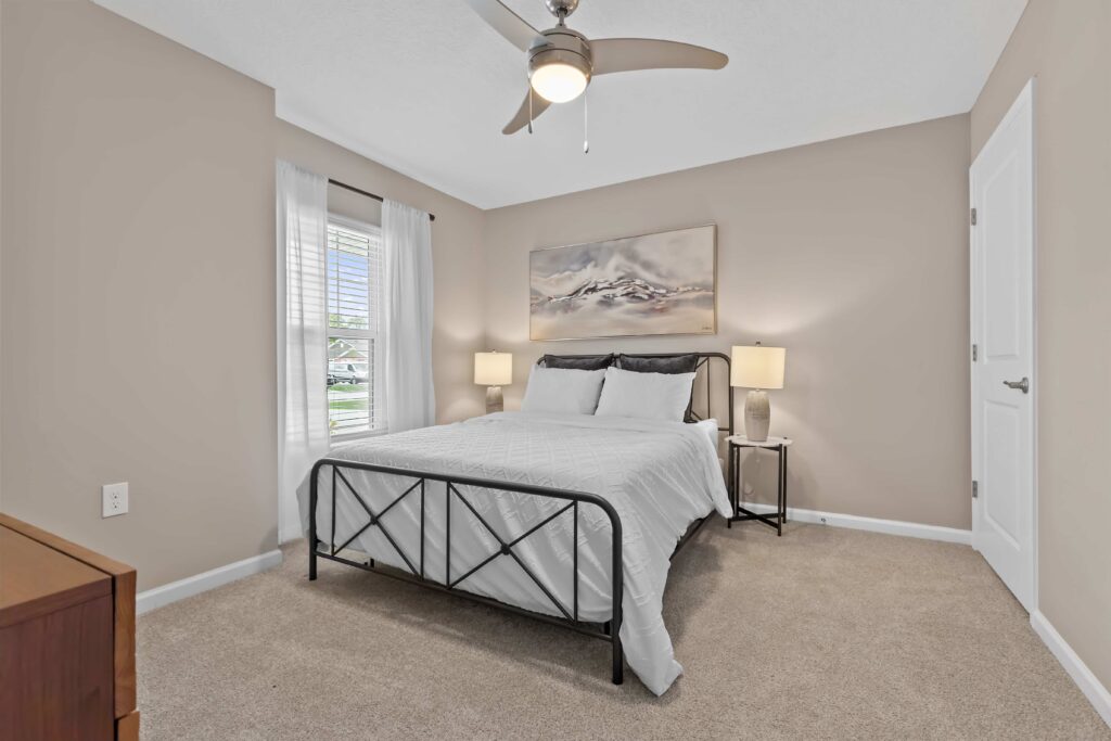 A neatly made bed with white bedding in a carpeted bedroom, flanked by two lamps on side tables, with a ceiling fan and a landscape painting above the bed.