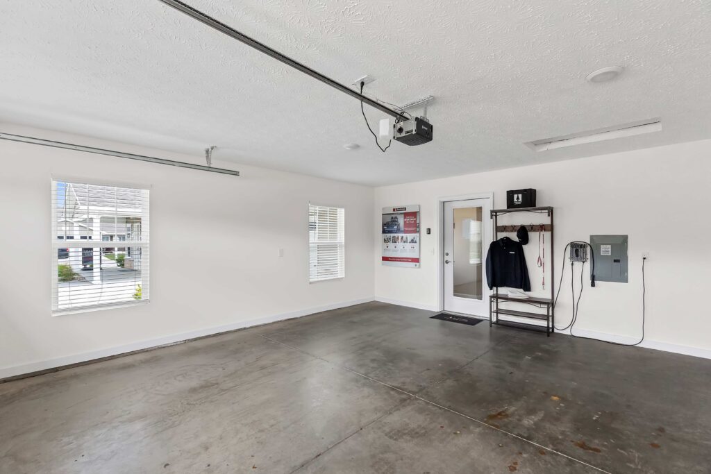Clean, empty garage with concrete floor, white walls, two windows, a door, wall-mounted controls, a jacket hanging, and a few items on a shelf.