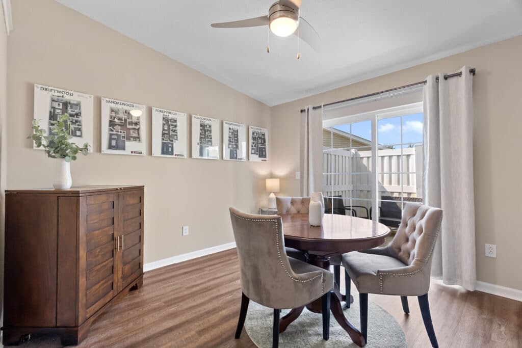 A round dining table with four tufted chairs sits near a window with white curtains; a cabinet and framed art decorate the beige-walled room with wooden flooring.
