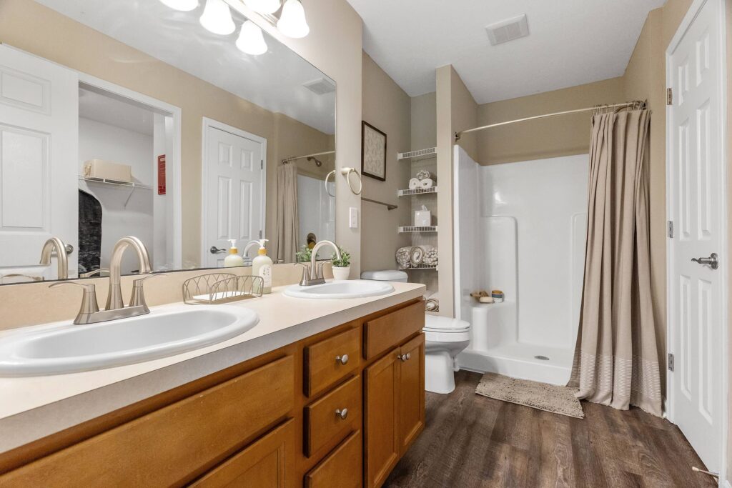 A bathroom with double sinks, a large mirror, wooden cabinets, open shelving with towels, a toilet, and a walk-in shower with a curtain.