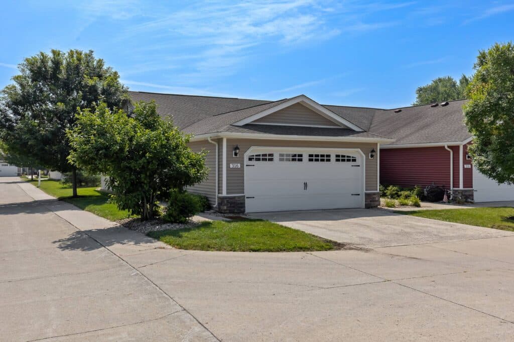 Single-story suburban house with beige siding, a double garage door, small front lawn, and trees, set along a paved street on a clear, sunny day.