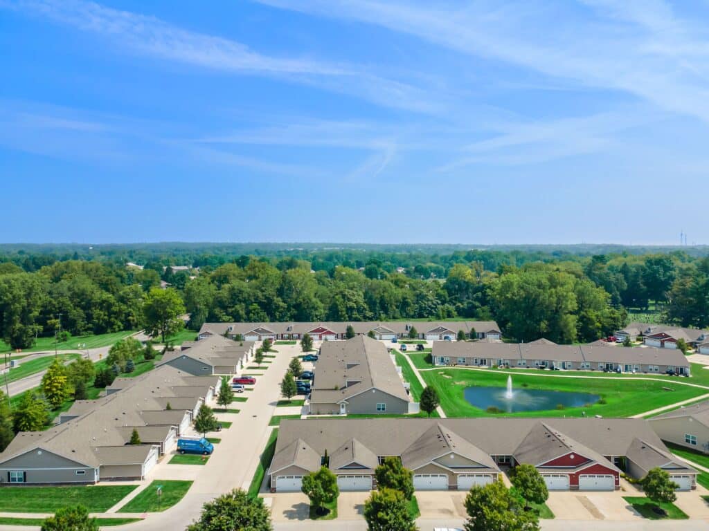 Aerial view of a suburban neighborhood with rows of houses, green lawns, trees, parked cars, and a small pond with a fountain under a blue sky.