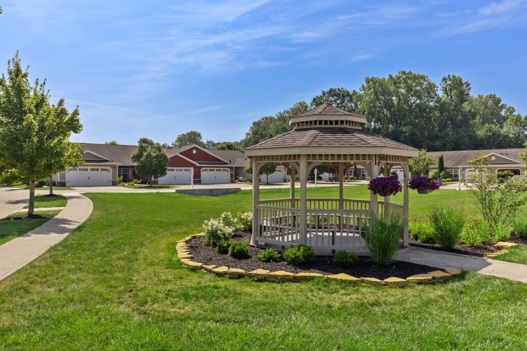A wooden gazebo stands on a landscaped lawn with flowers, surrounded by sidewalks and single-story houses under a blue sky.