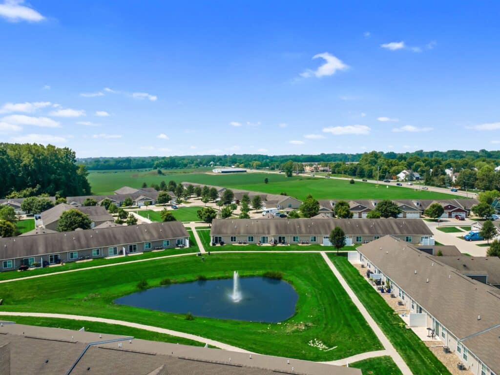 Aerial view of a residential complex with single-story buildings, a central pond with a fountain, sidewalks, green lawns, and trees, surrounded by open fields and rural landscape.