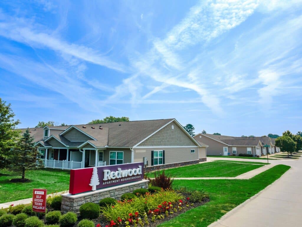 Single-story apartment buildings with manicured lawns and a sign reading "Redwood Apartment Neighborhoods" under a blue sky with wispy clouds.