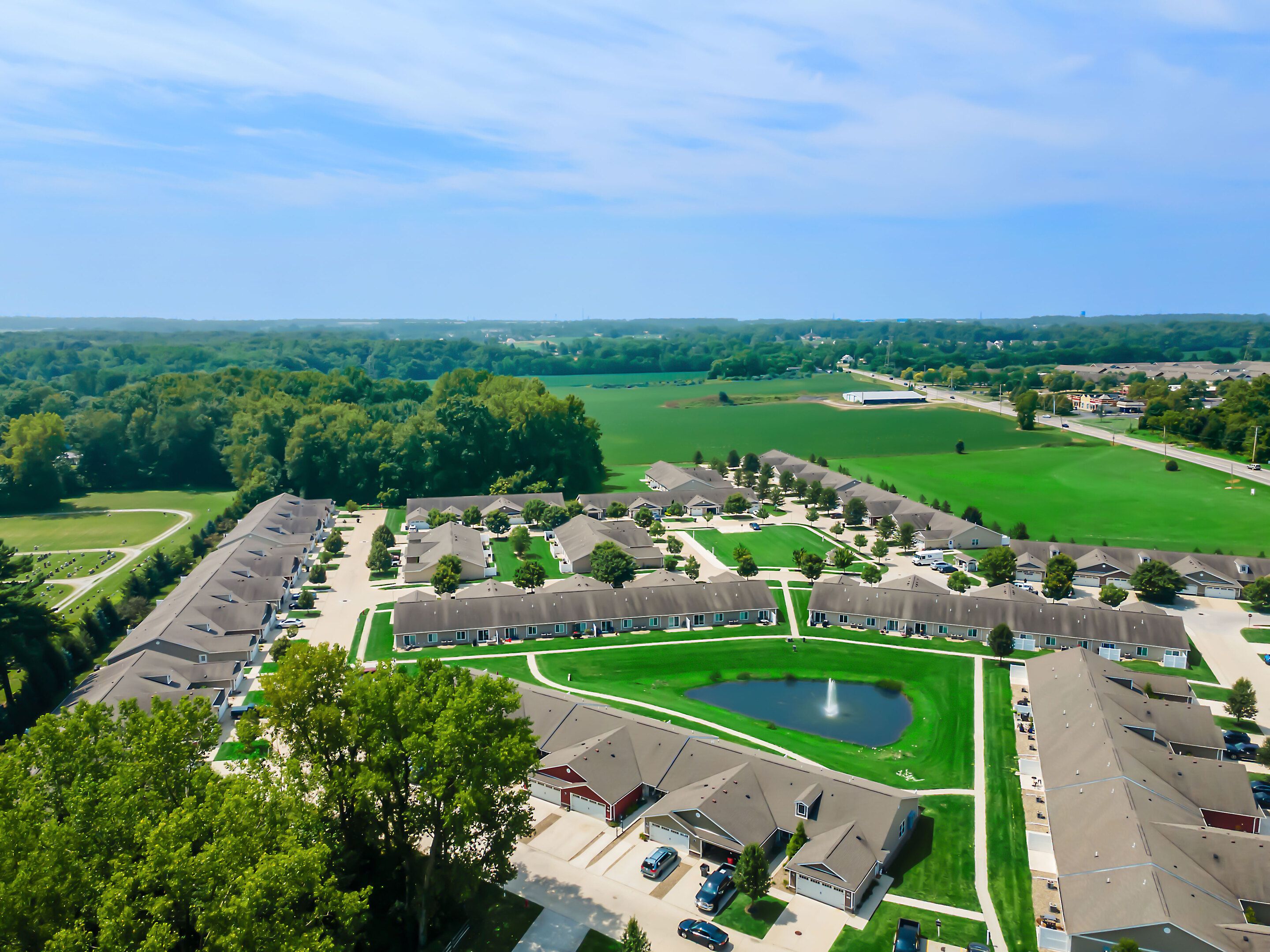 Aerial view of a residential neighborhood with rows of houses, a small pond with a fountain, and surrounding green fields under a blue sky.