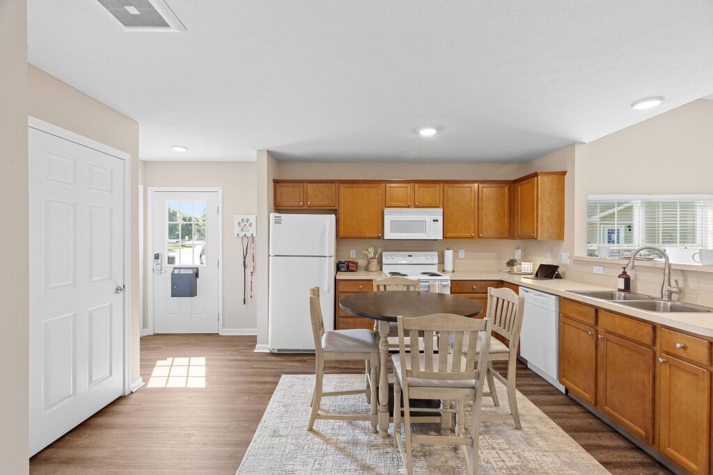 A kitchen with wooden cabinets, white appliances, a round dining table with four chairs, and a window above the sink. The floor is wood and a rug is under the table.