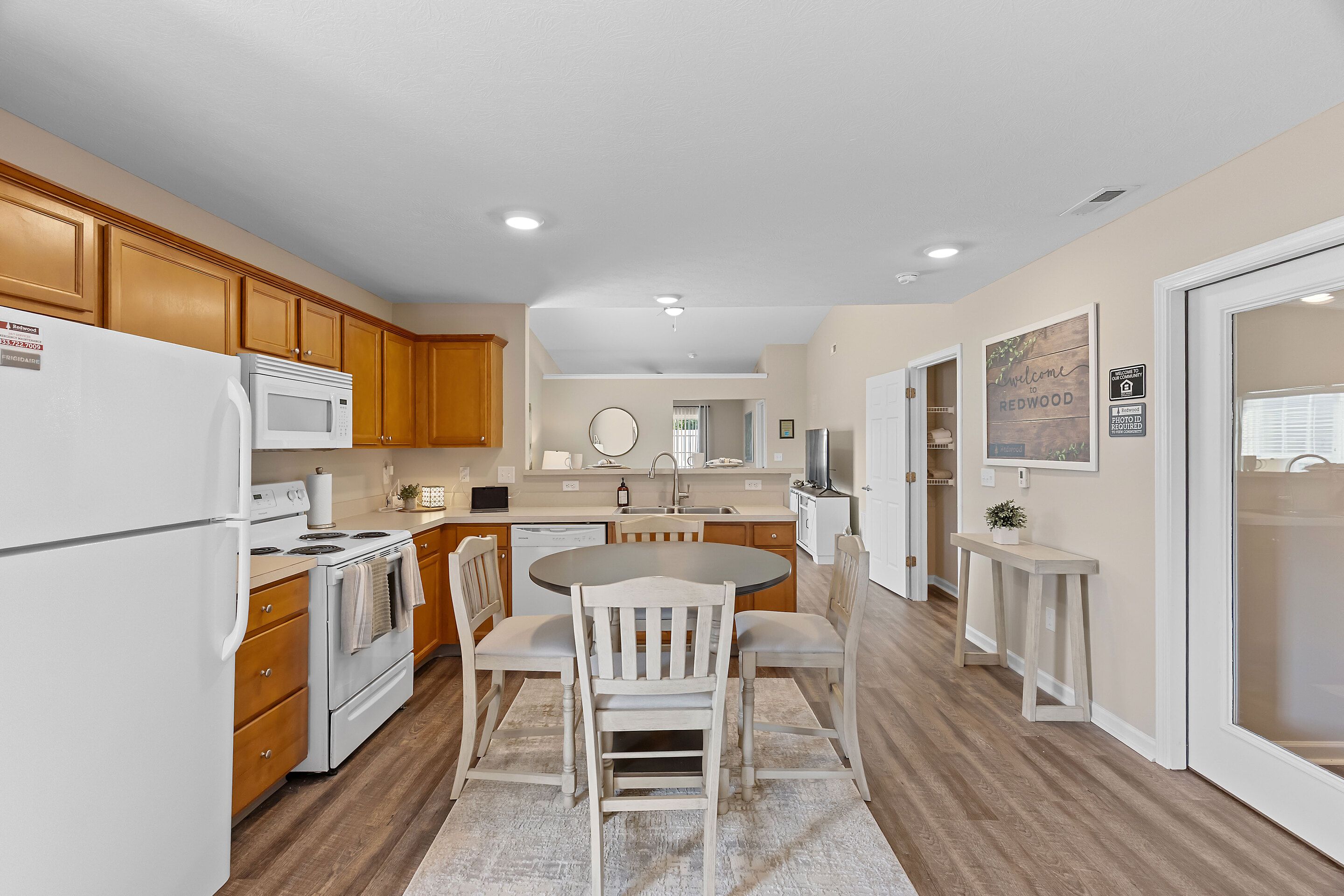 A kitchen and dining area with wood cabinets, white appliances, a round table with four chairs, and neutral walls with framed artwork.