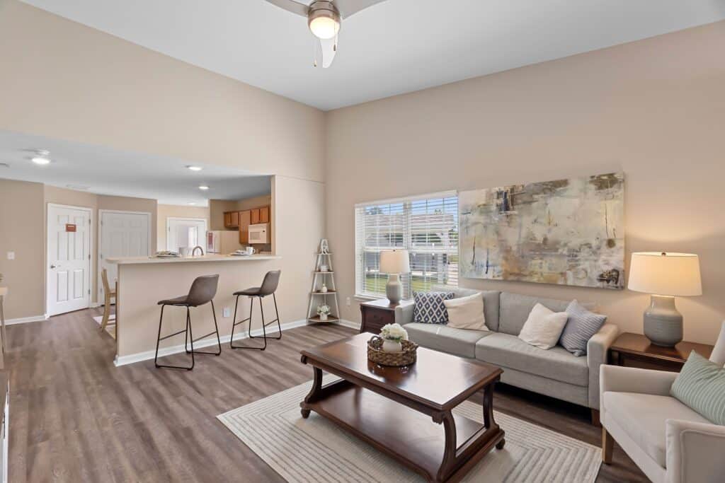 A cozy living room with a sofa, coffee table, and lamp, adjacent to a kitchen with barstools and an open layout. Neutral colors and modern decor.