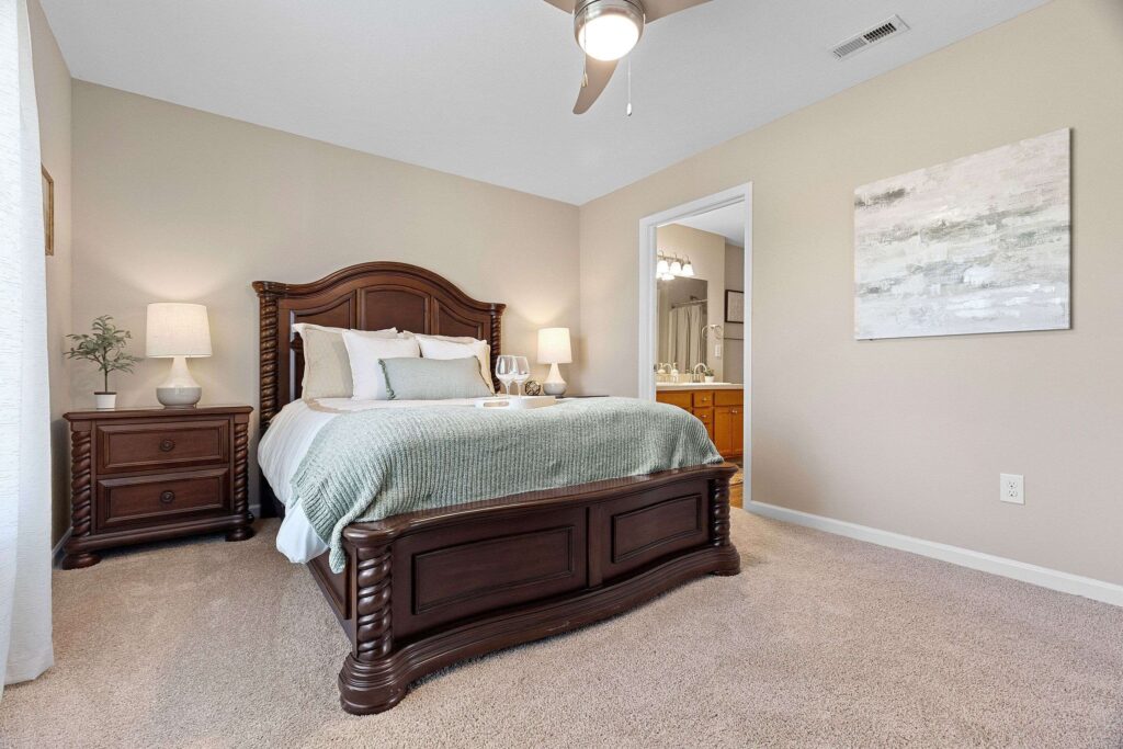 A bedroom with a wooden bed frame, matching nightstands, table lamps, neutral walls, carpeted floor, and an open doorway to a bathroom.