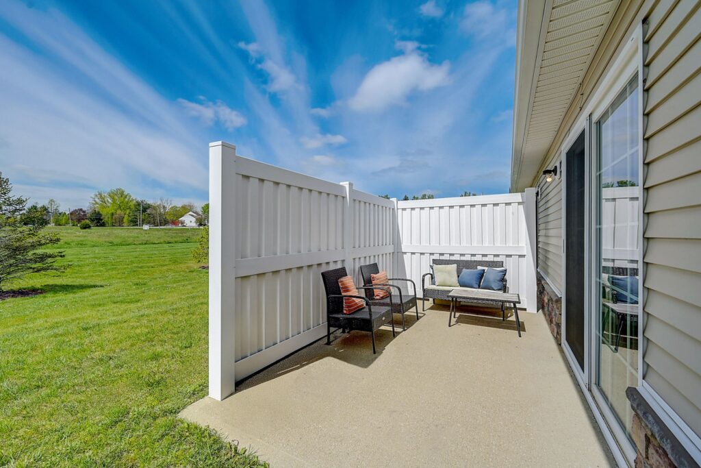 Small patio with two chairs, a bench with cushions, and a white privacy fence next to a beige house; grassy yard and blue sky in the background.
