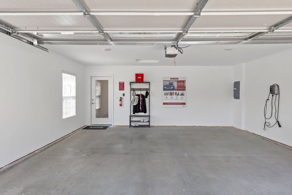 Empty two-car garage with white walls, exposed ceiling tracks, storage shelves, wall-mounted equipment, a window, and a door to the interior.