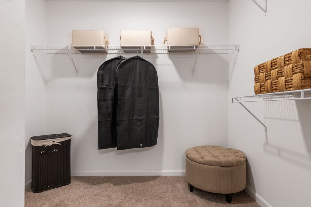 A tidy walk-in closet with wire shelves holding boxes, a black garment bag, a round beige ottoman, a basket, and a hamper.