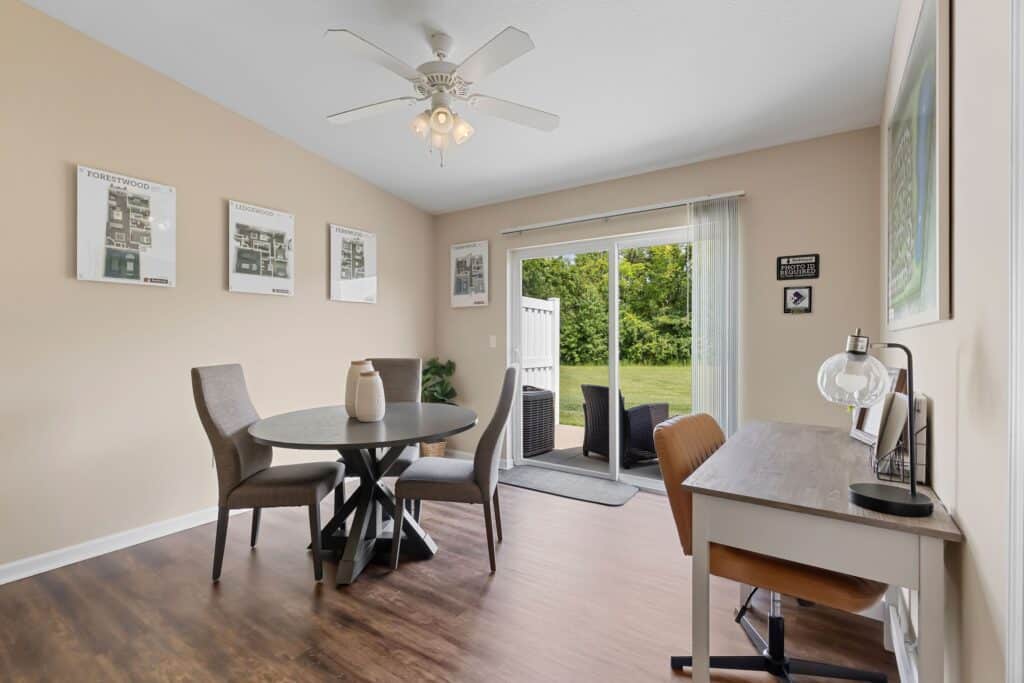 A dining area with a round table and four chairs, adjacent to a desk with a lamp, near glass doors that open to a patio and green outdoor space.