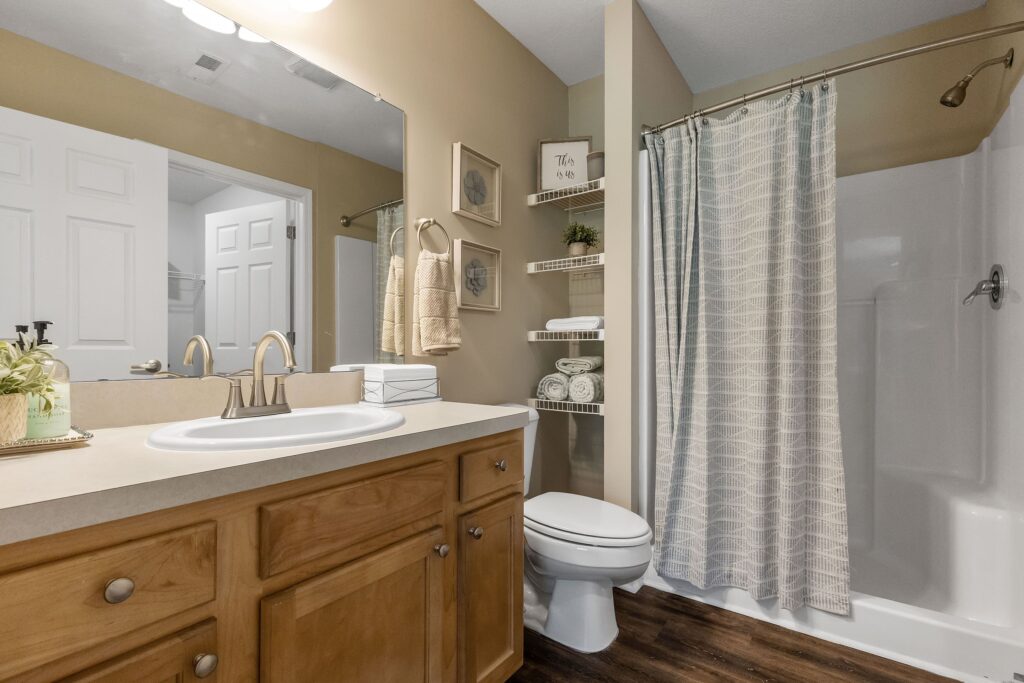 A bathroom with a wood vanity, white sink, toilet, open shelves with towels, and a shower with a curtain; neutral tones and simple decor.
