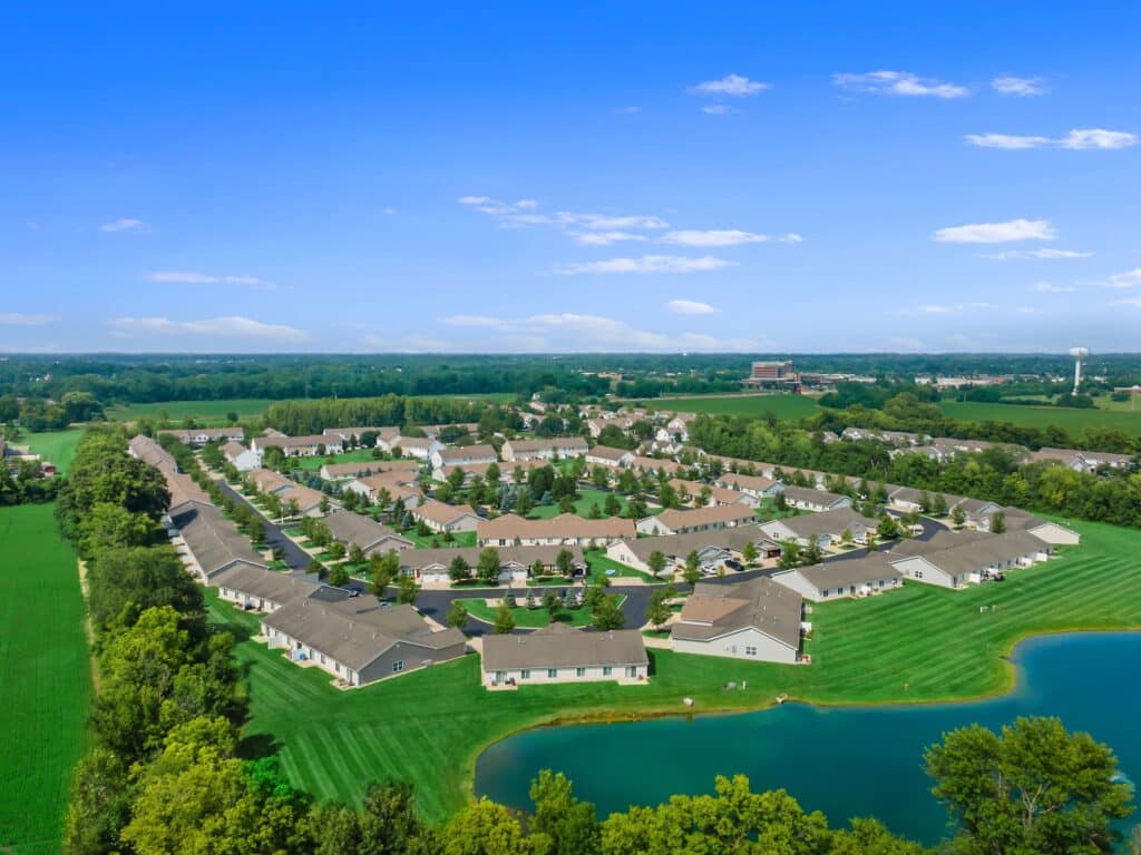Aerial view of a suburban neighborhood with circular streets, houses, green lawns, trees, and a small pond, surrounded by open fields and farmland under a clear blue sky.