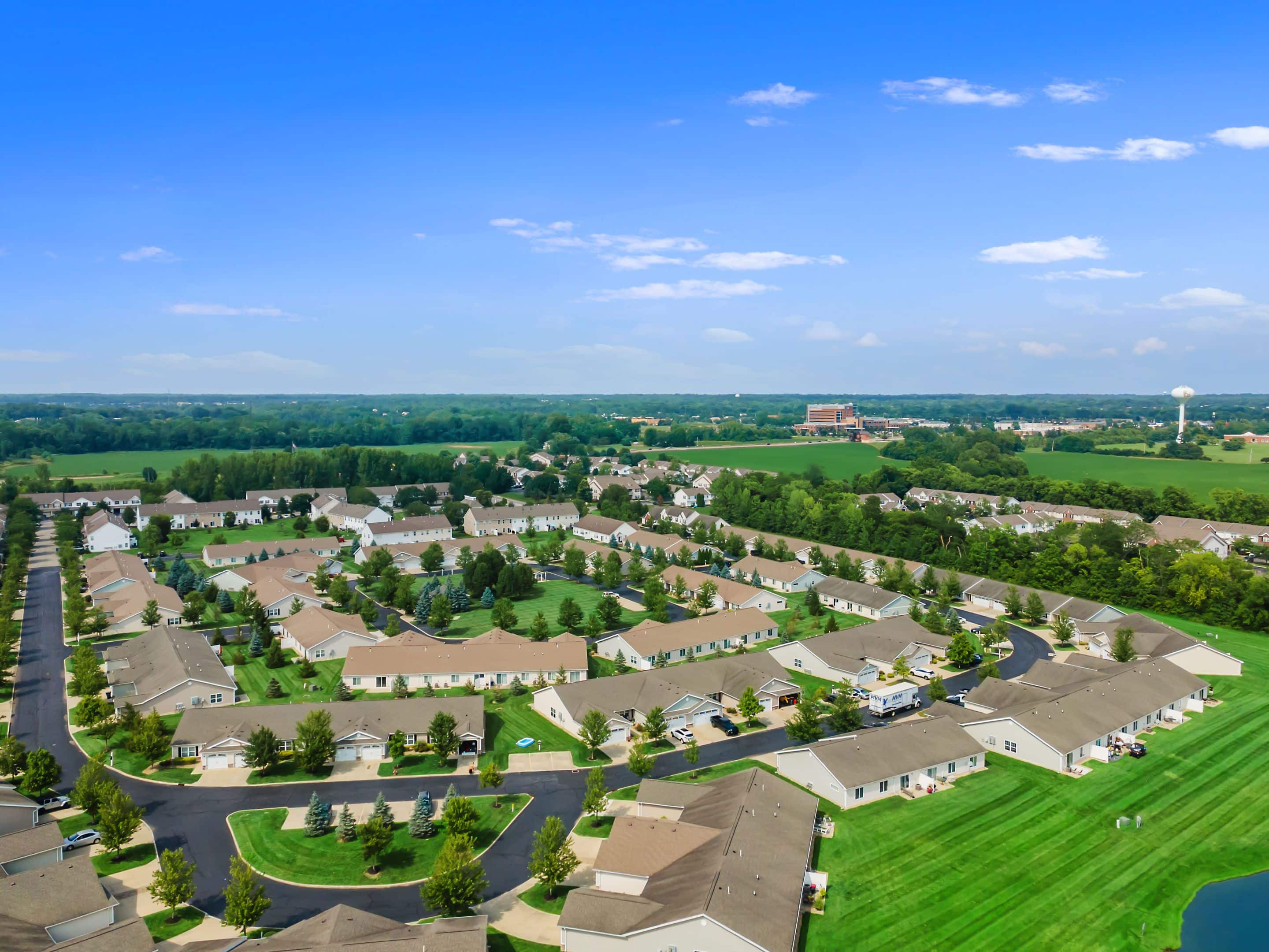 Aerial view of a suburban residential neighborhood with rows of houses, green lawns, trees, and surrounding open fields under a blue sky.