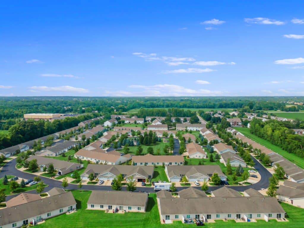 Aerial view of a suburban residential neighborhood with rows of single-story houses, green lawns, and trees under a blue sky.