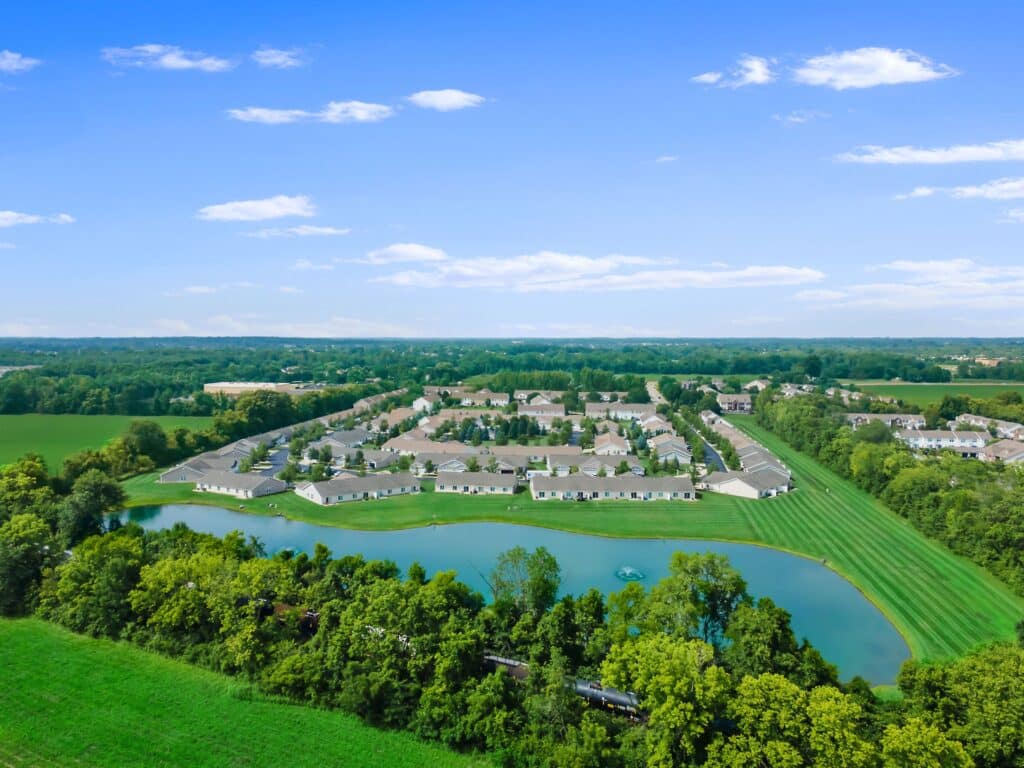 Aerial view of a residential community surrounded by green fields and trees, with a pond in the foreground under a clear blue sky.