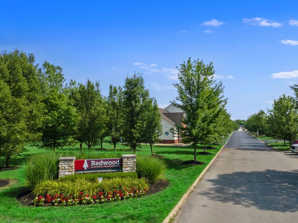 A street view shows a landscaped entry sign for Redwood apartment homes surrounded by green trees and grass on a clear, sunny day.