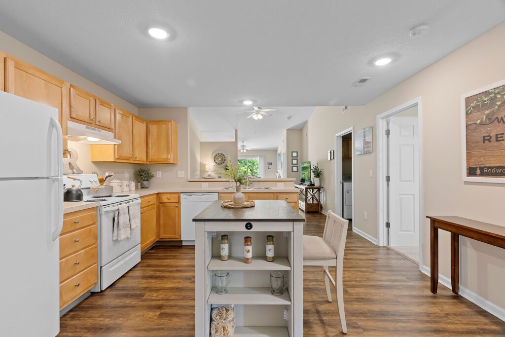 A modern kitchen with light wood cabinets, white appliances, a small island, and wood flooring, opening into a bright living area with windows and greenery visible outside.