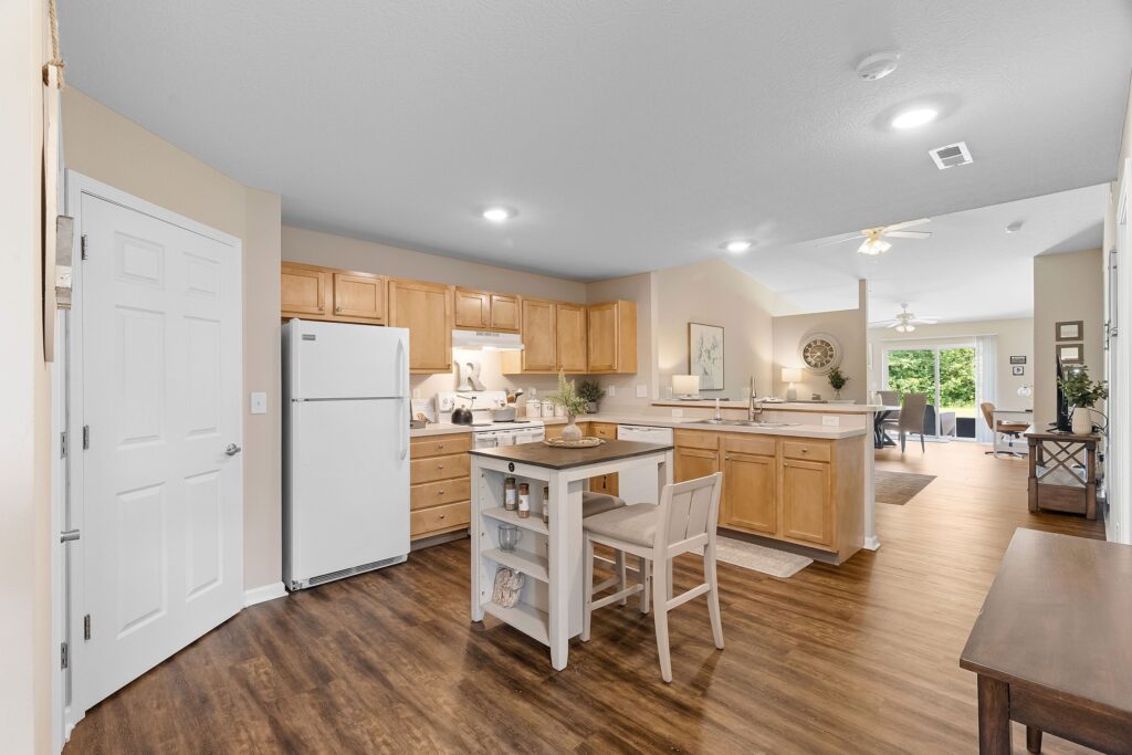 A modern kitchen with light wood cabinets, white appliances, an island with chairs, and wood flooring, opening to a dining and living area with large windows.