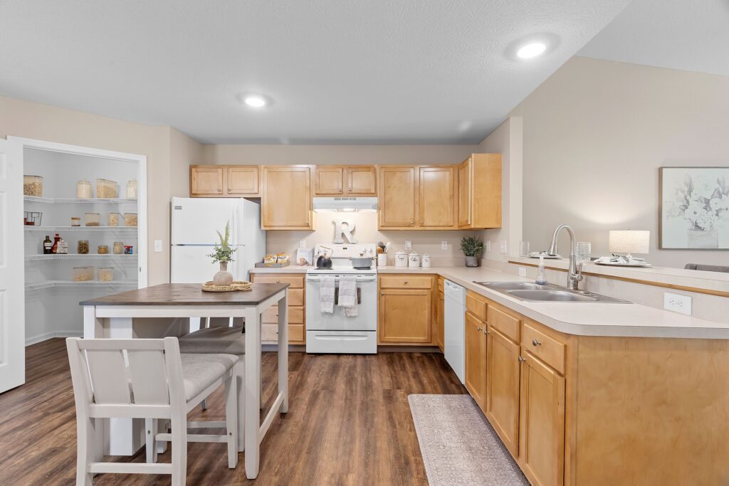 Bright kitchen with light wood cabinets, white appliances, a small dining table with two chairs, and a walk-in pantry with shelves filled with jars and baskets.