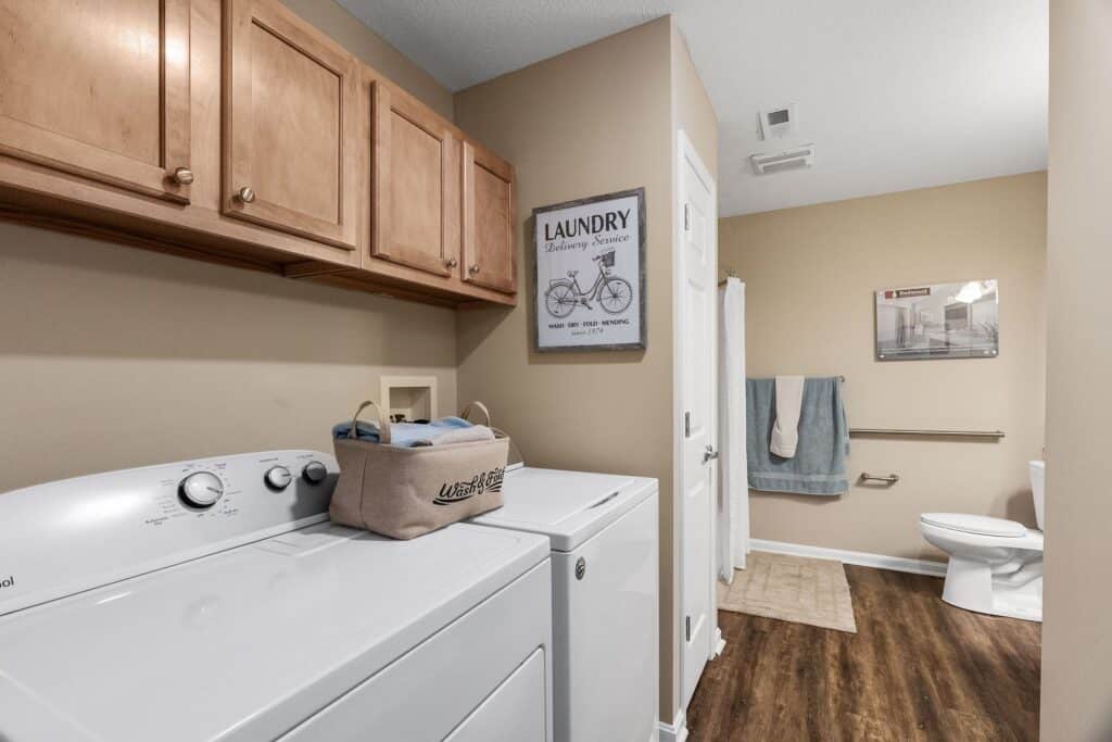 A laundry room with a washer and dryer, wooden cabinets, a laundry basket, and wall-mounted decor, leading into a bathroom with a toilet and towel racks.