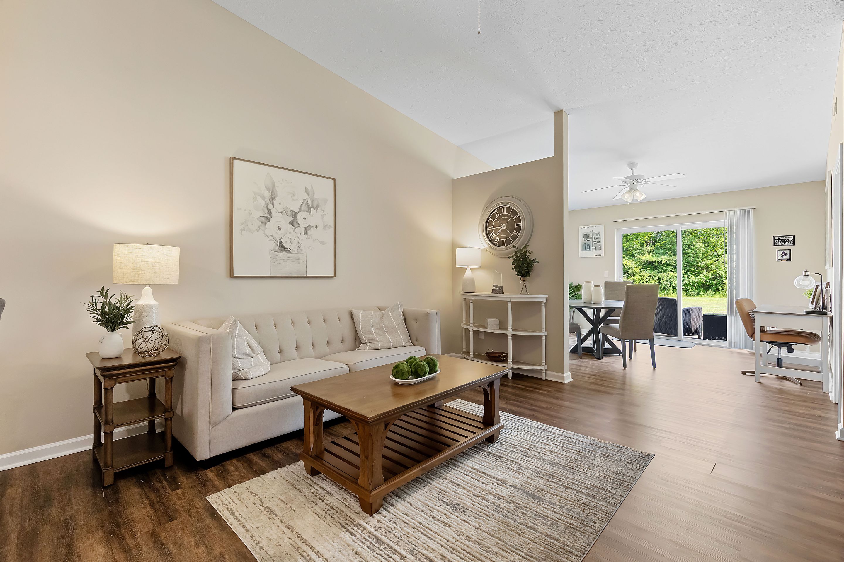 A bright living room with a beige sofa, wooden coffee table, and wall art, leading to a dining area with large windows overlooking greenery.