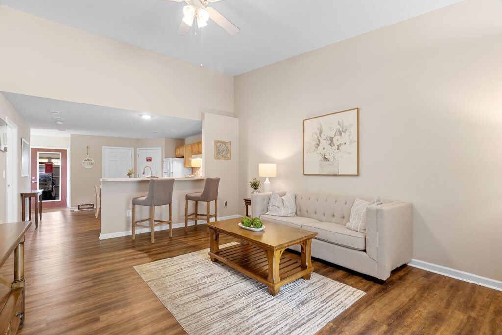 A living room with a beige sofa, a wooden coffee table on a rug, and an open kitchen with two bar stools and white cabinets in the background.