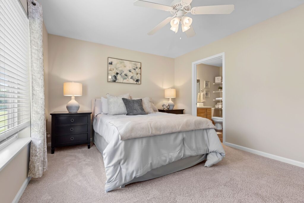 Bedroom with a neatly made bed, two nightstands with lamps, a ceiling fan, wall art above the bed, and an attached bathroom visible through an open door.