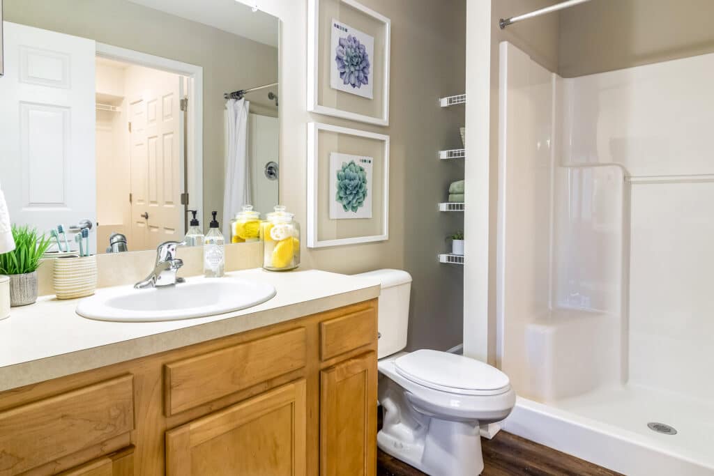 A bathroom with a wooden vanity, sink, toilet, walk-in shower, wall art, shelves with towels, and soap and lemon jars on the countertop.