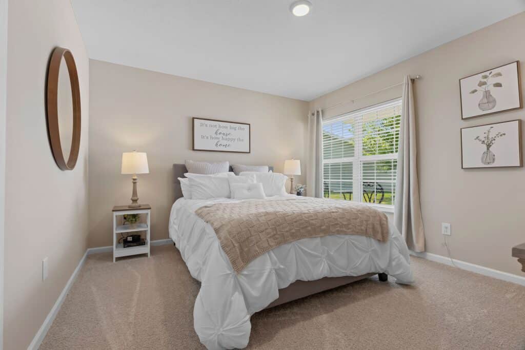 A neatly made bed with white bedding, beige blanket, and pillows is centered in a carpeted bedroom with two lamps, a round mirror, and two framed floral prints on the wall.