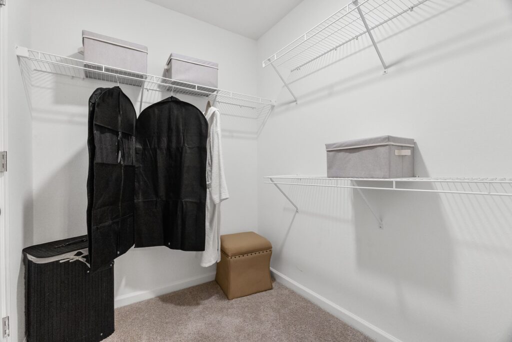 Empty walk-in closet with wire shelving, a laundry basket, two garment bags, fabric storage boxes, and a tan ottoman on carpeted floor.