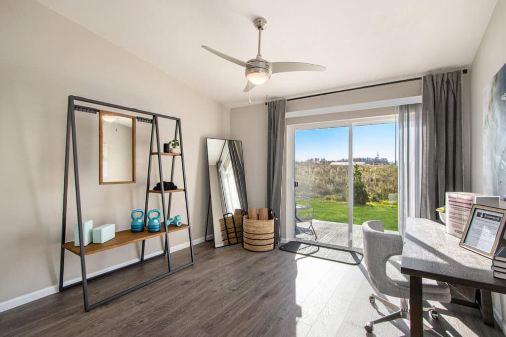 Modern home office with exercise equipment, a desk, shelving, floor mirror, and sliding glass doors opening to a green outdoor area.