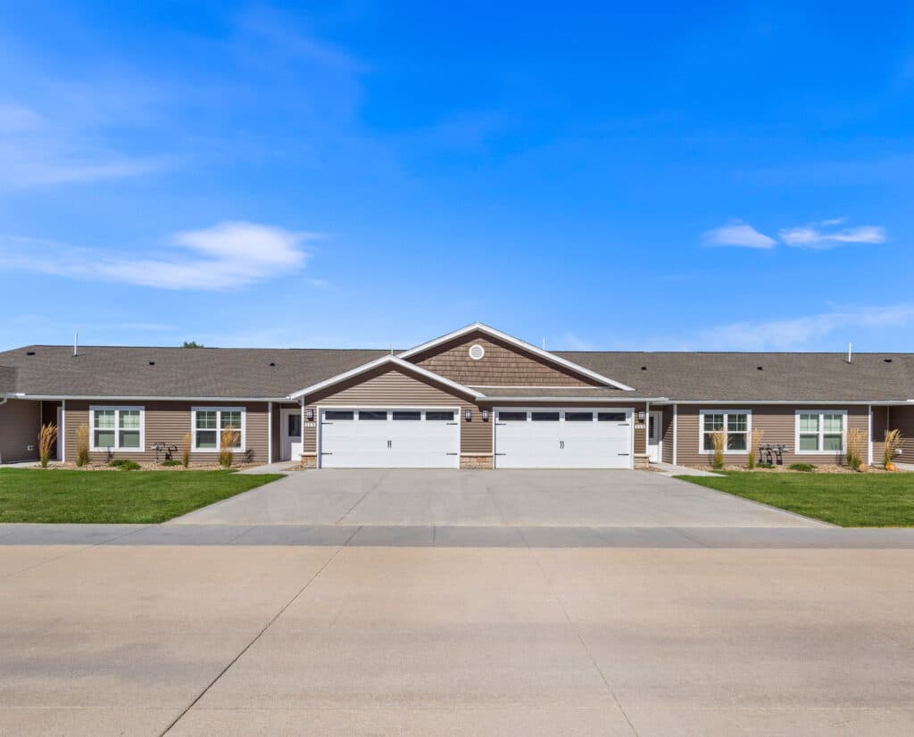 Single-story duplex building with two double garages, brown siding, white trim, and a wide concrete driveway under a clear blue sky.