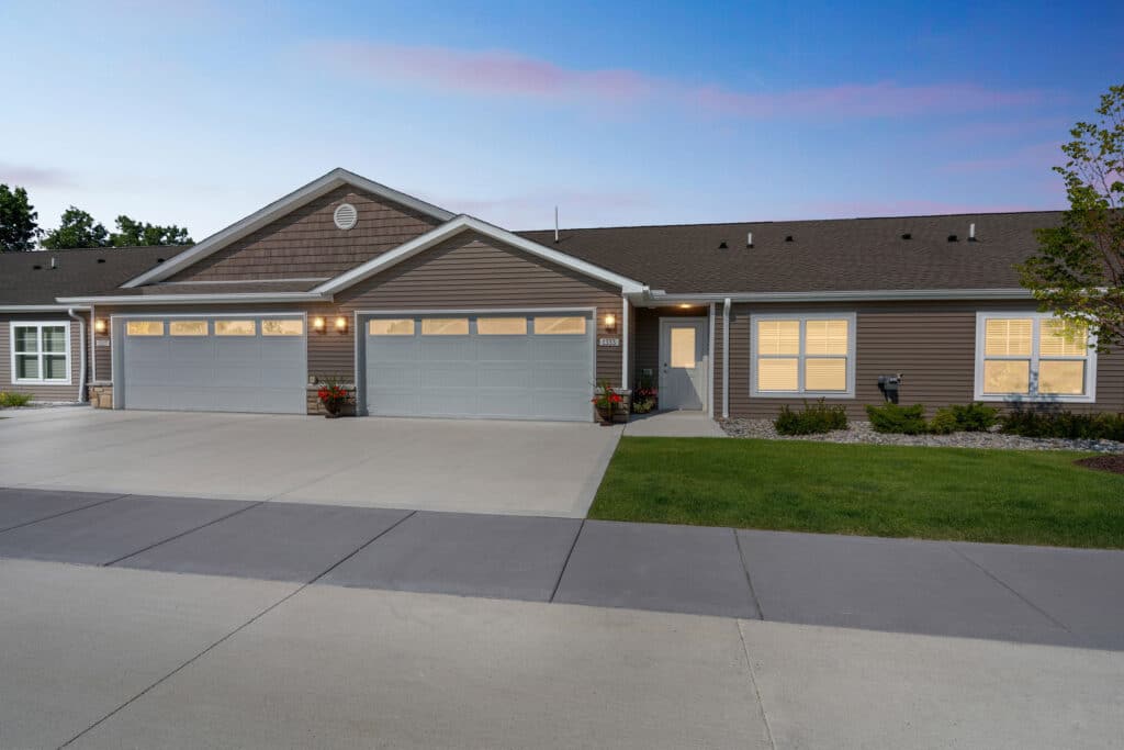 Single-story duplex with two double garages, brown siding, and well-maintained lawns, photographed at dusk with lights on inside the home.