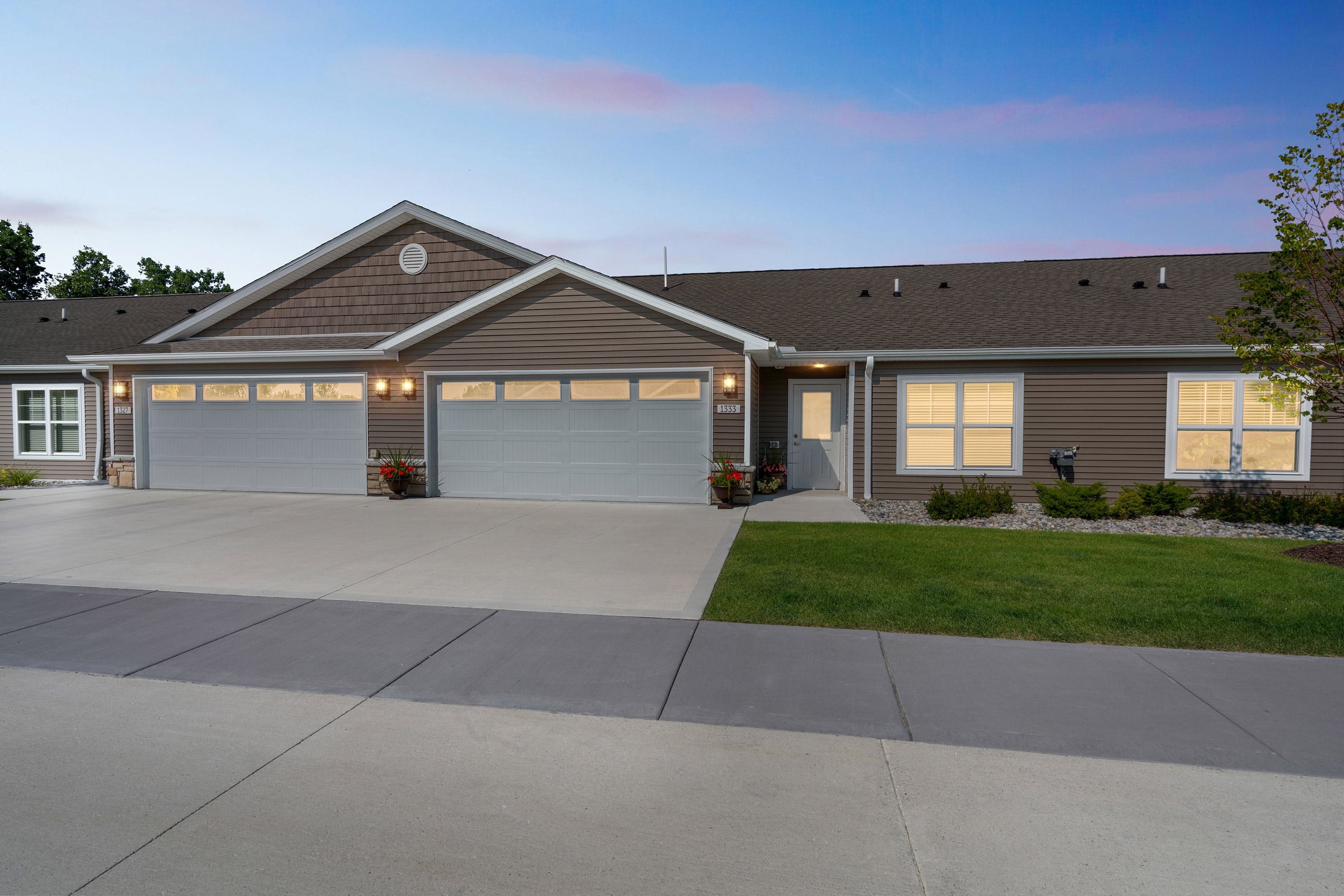 Single-story duplex with two double garages, brown siding, and well-maintained lawns, photographed at dusk with lights on inside the home.